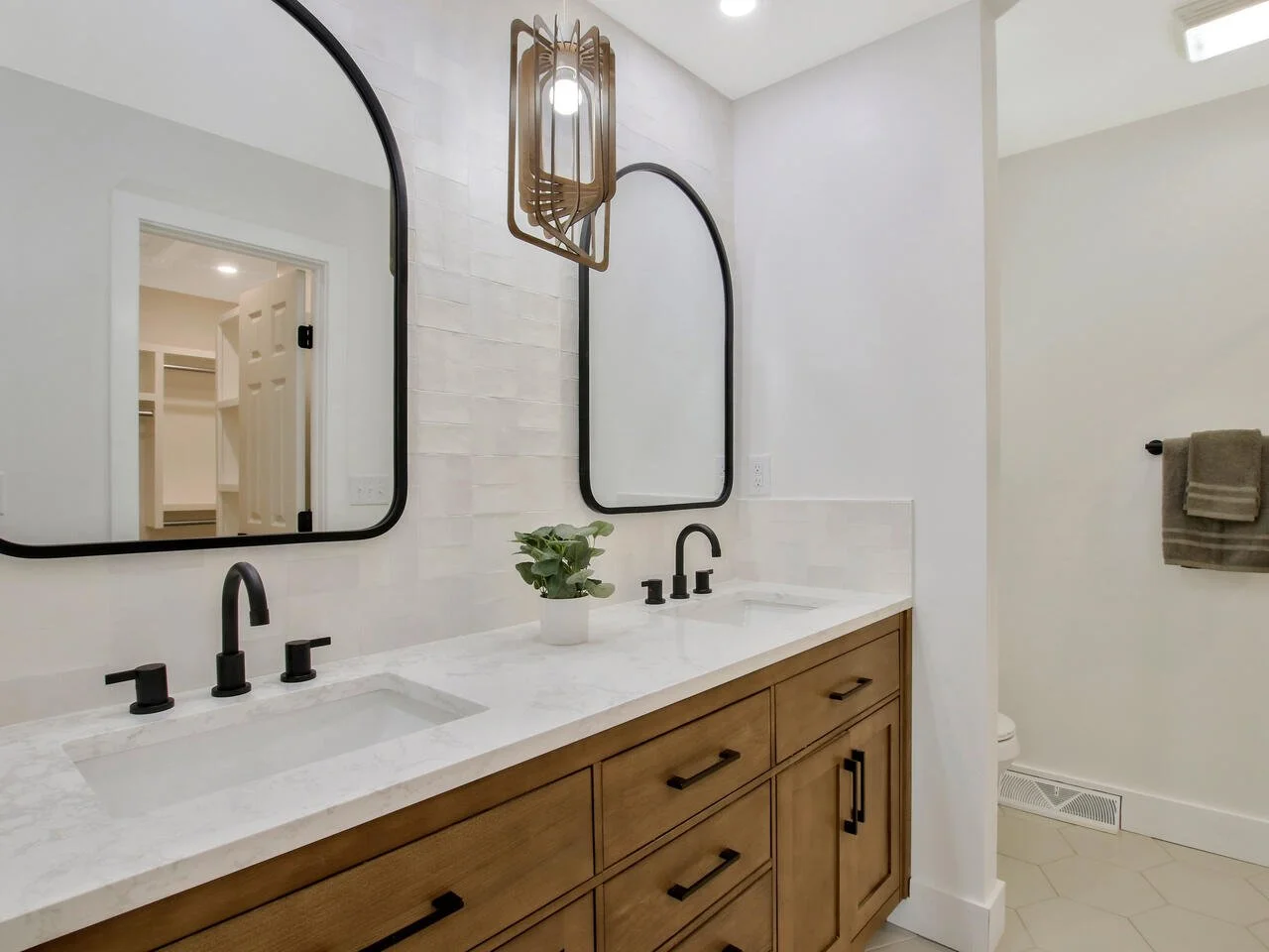 Modern bathroom vanity with two sinks, black faucets, wooden cabinets, mirrored frames, a small potted plant, and a hanging light fixture.