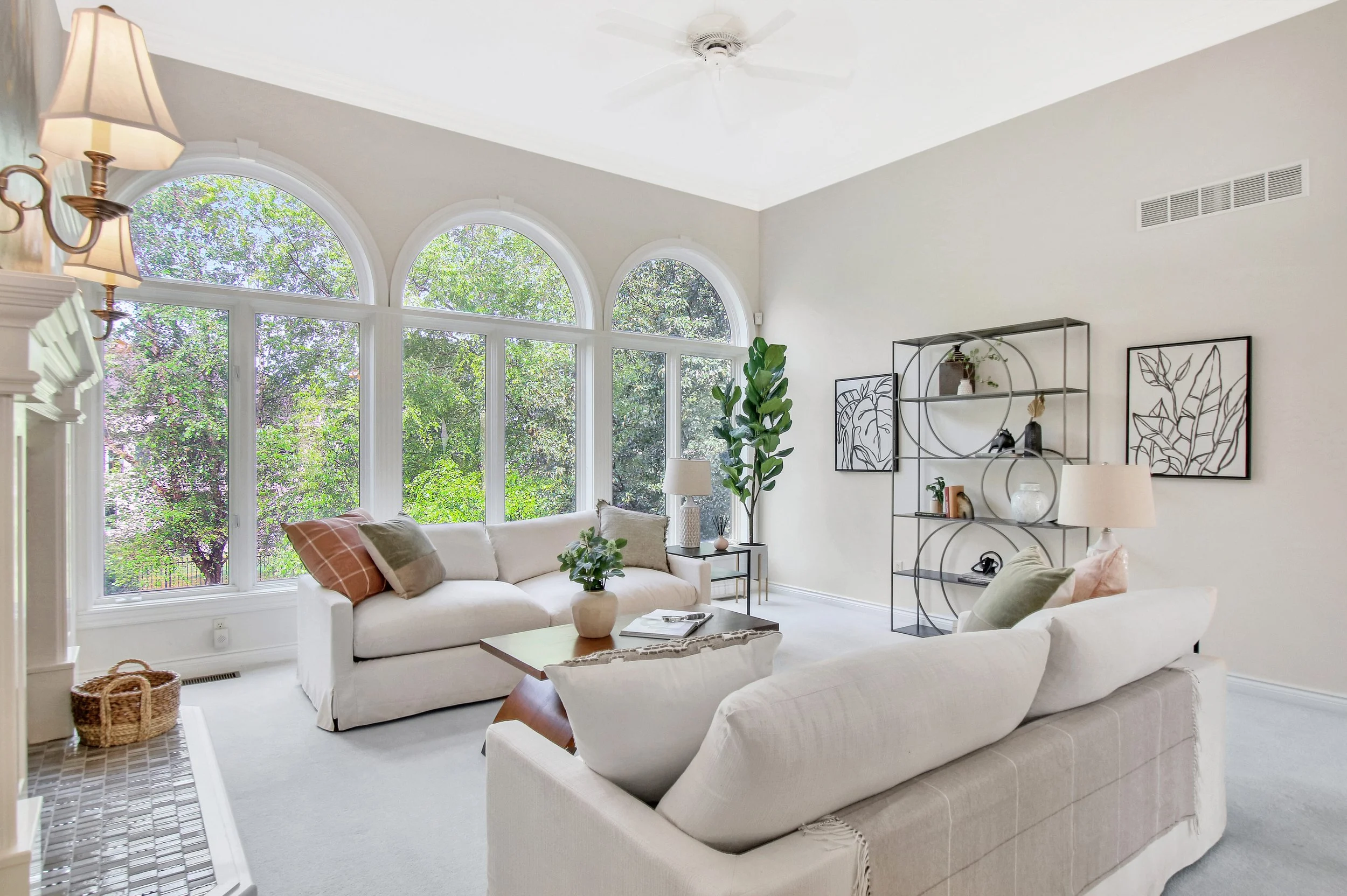 Bright living room with large arched windows, white sofas, a gray and white armchair, a tall green plant, decorative shelving, framed leaf artwork, and natural light.