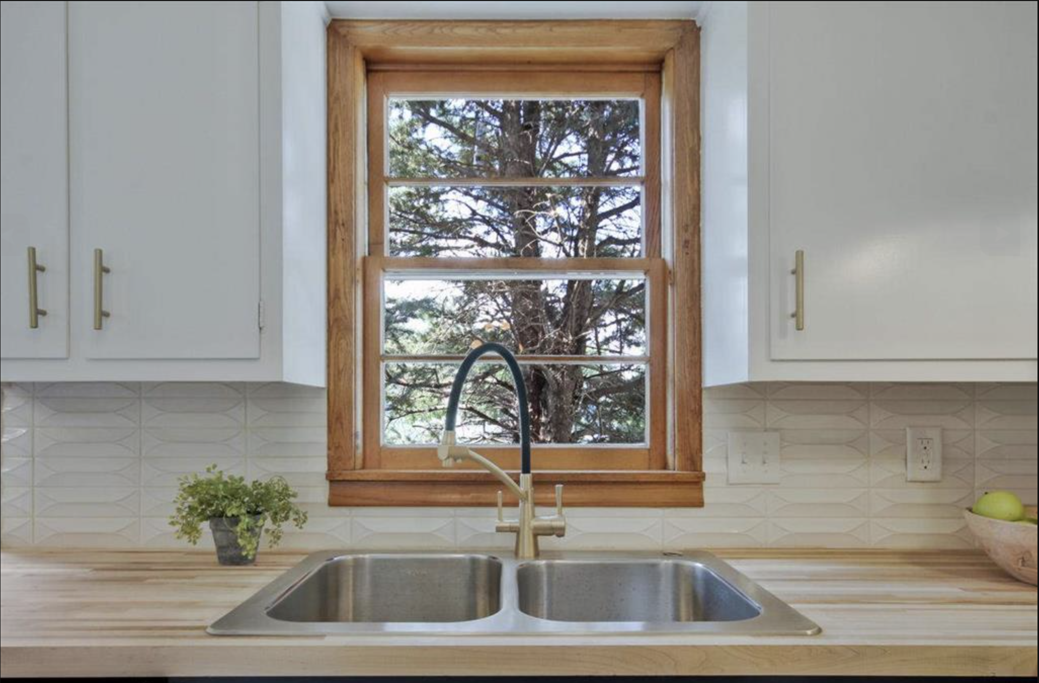Kitchen sink with a window showing outdoor trees, white cabinets, wooden countertop, a small potted plant on the left, and a bowl with bananas on the right.