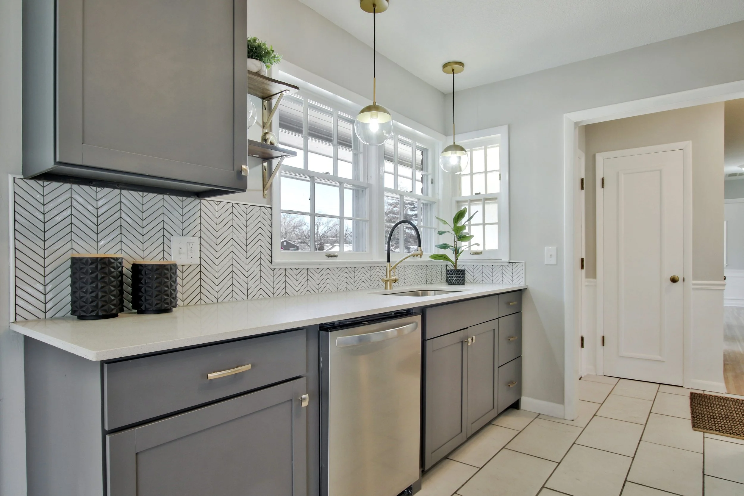 Modern kitchen with grey cabinets, white countertop, and patterned tile backsplash. Large window behind the sink with sunlight, decorative plants, and two hanging pendant lights.