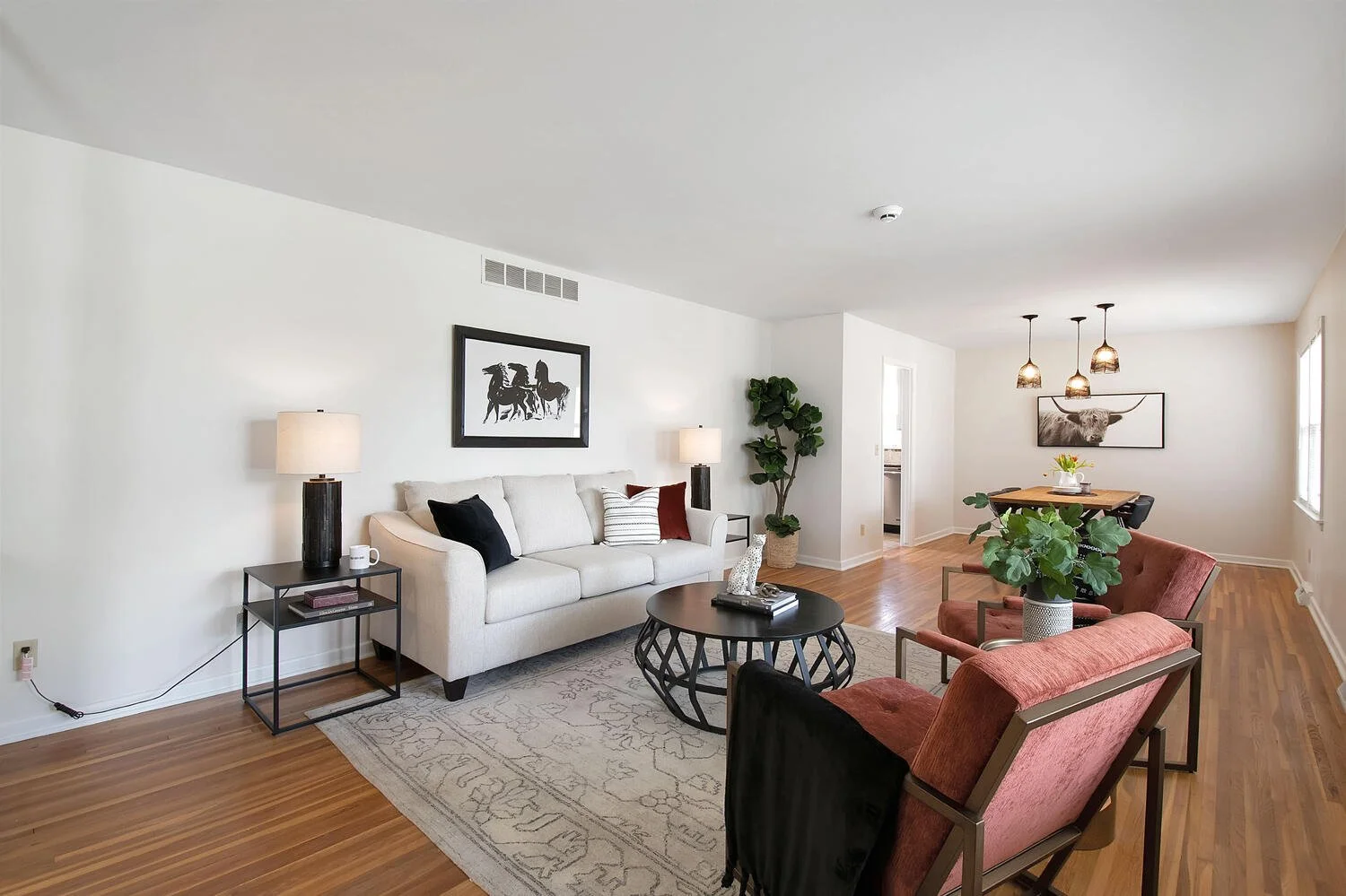 Living room with white couch, black side table, beige rug, two lamps, black and rust-colored chairs, wall art, houseplant, and wooden dining table with pendant lights.