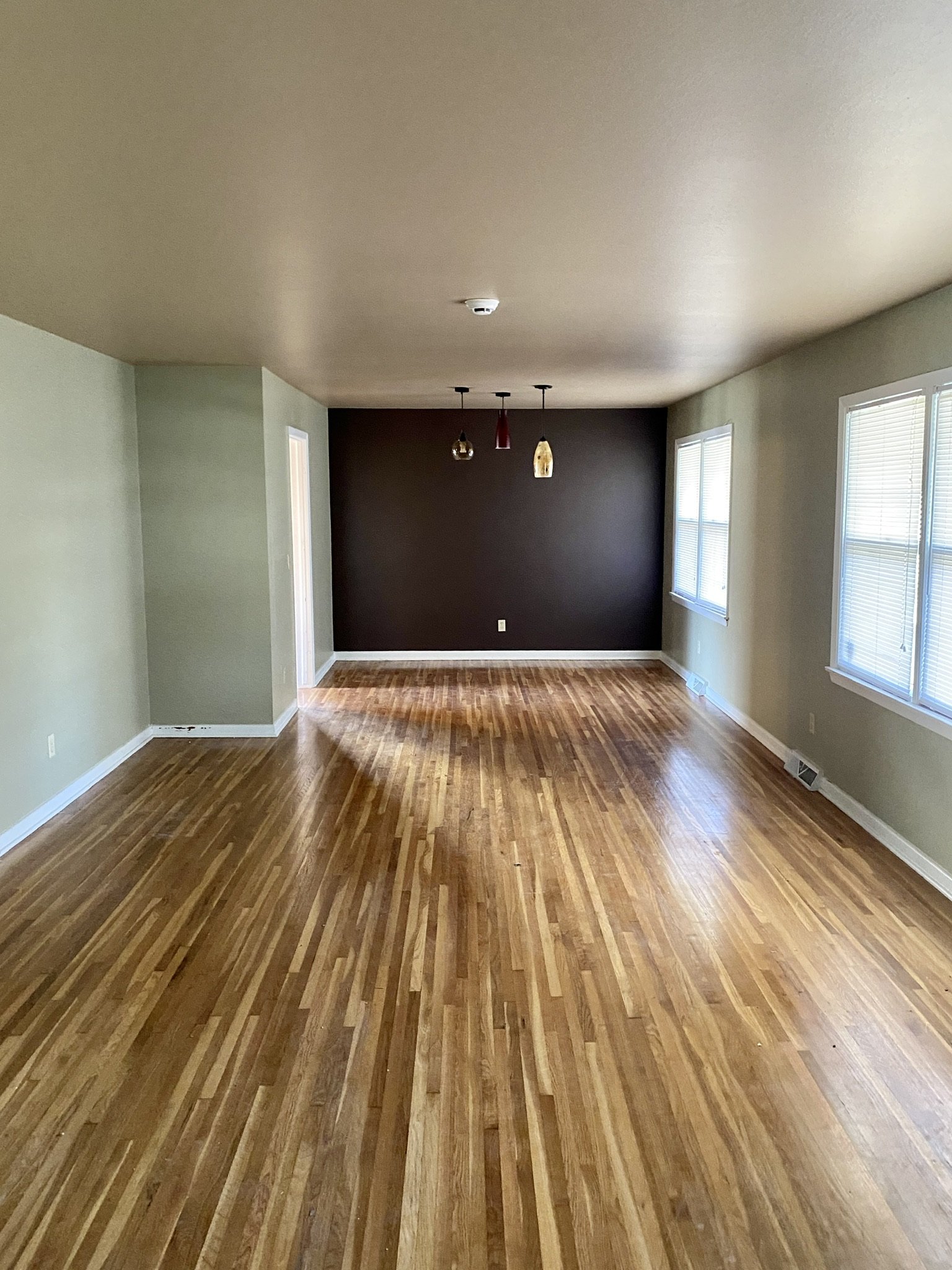 Empty living room with hardwood floors, three large windows with blinds, a dark accent wall with three hanging lights, and light-colored walls.