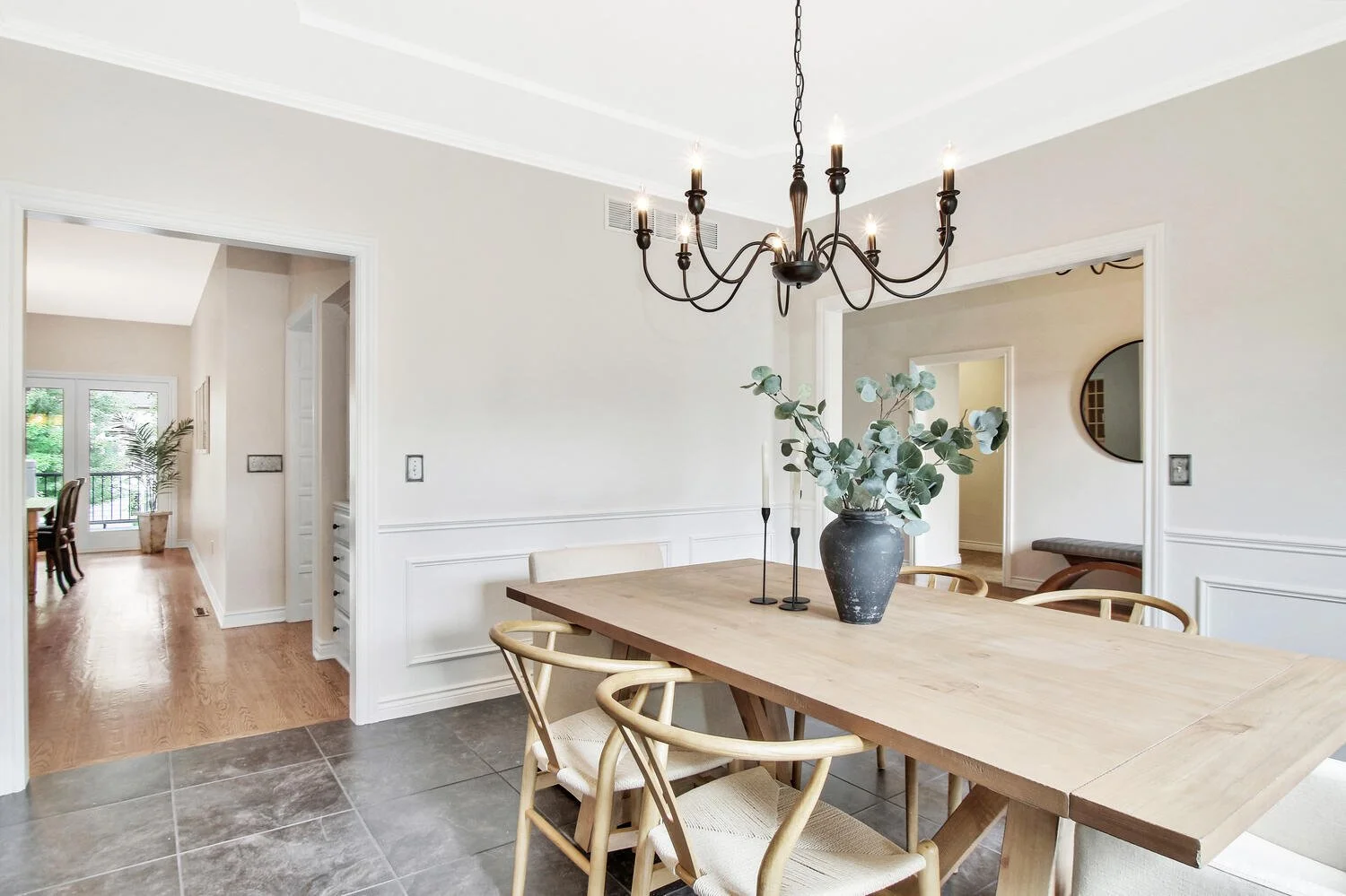 Dining room with a wooden table, six chairs, a black vase with eucalyptus, a black chandelier, and a doorway leading to a room with a large window, white walls, and hardwood flooring.