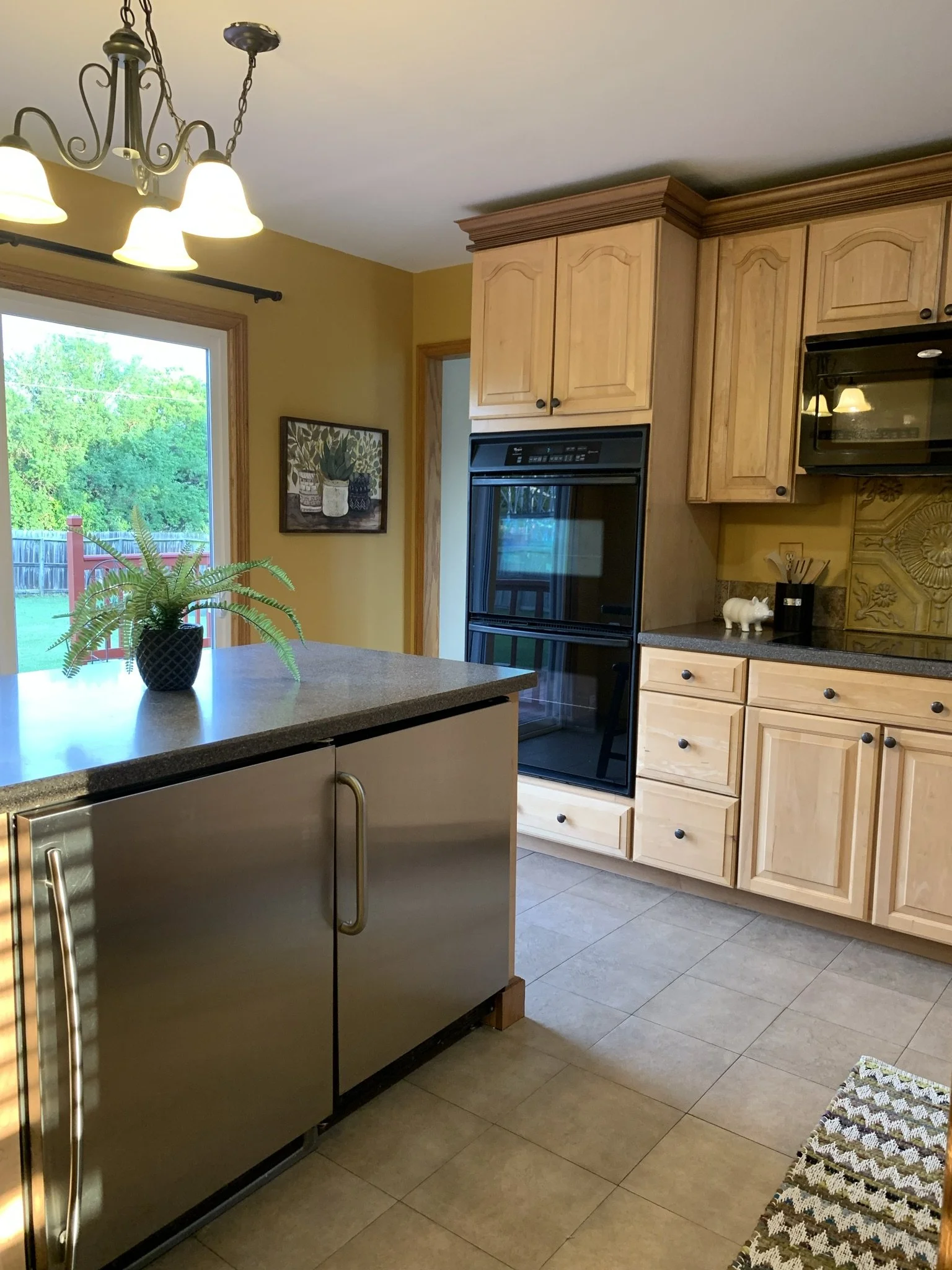 A kitchen with wooden cabinets, black appliances, a tiled floor, a window showing a backyard, and a kitchen island with a potted plant.