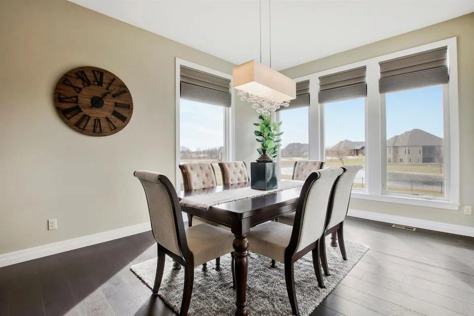 A dining room with a dark wood table and six beige upholstered chairs, a large wall clock, a modern chandelier, and large windows with blinds overlooking a suburban neighborhood.