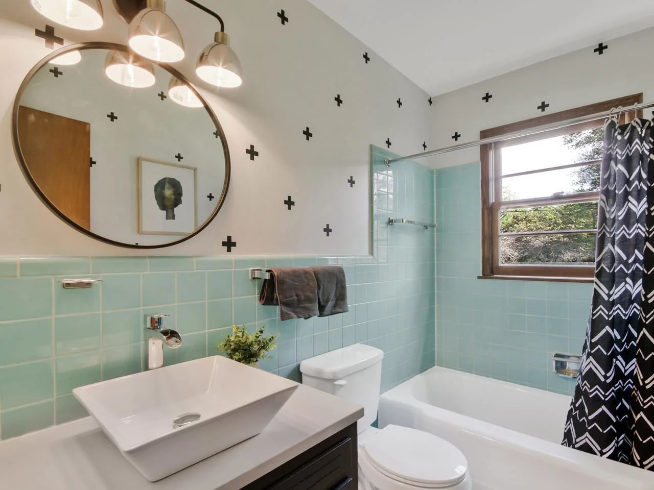 Bathroom with light green tiles, round mirror, modern sink, toilet, bathtub, window with wooden frame, patterned shower curtain, wall art, towel rack with dark towel, and ceiling lights.