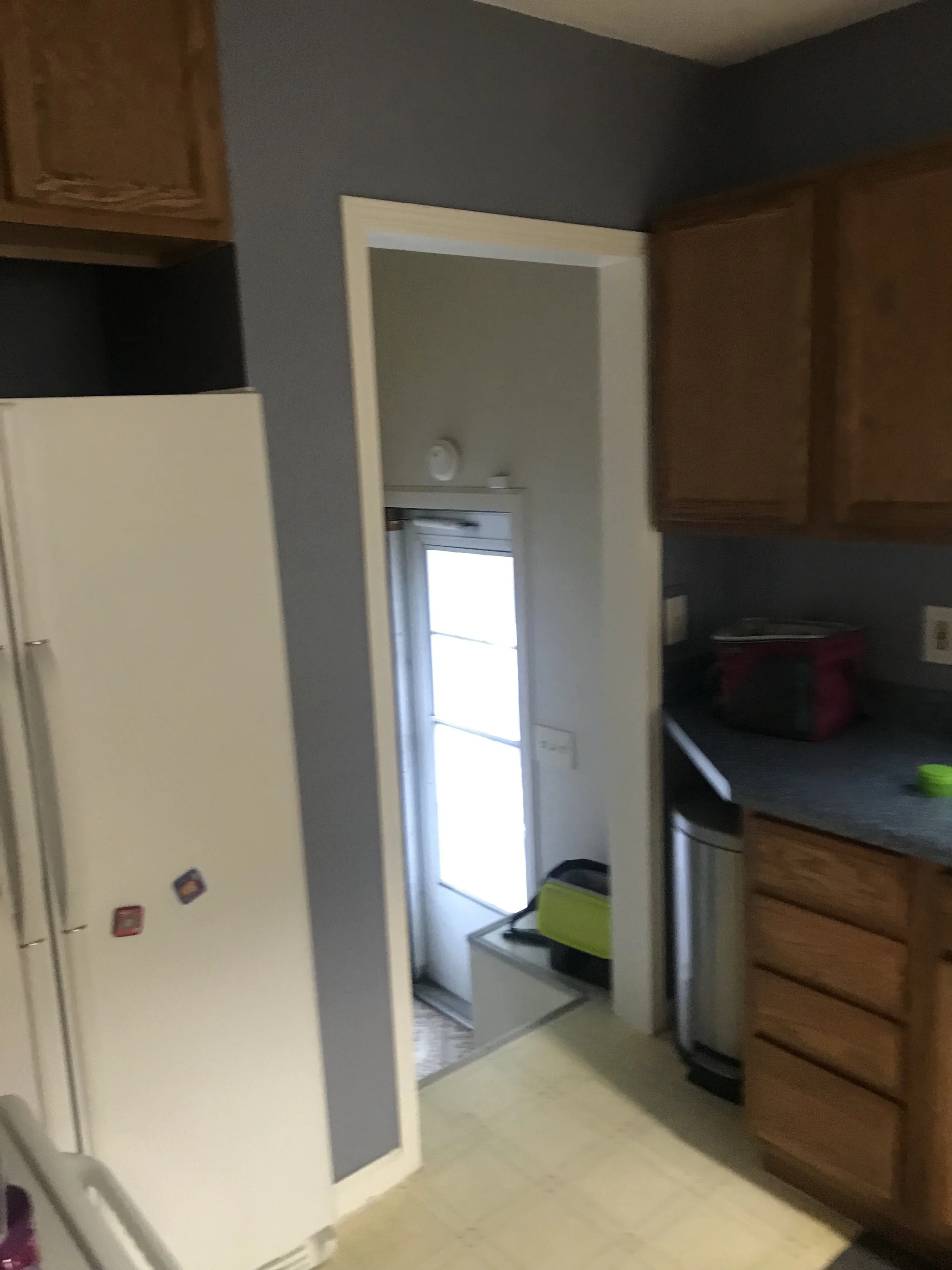 View of a kitchen with a door leading outside, wooden cabinets, a countertop with a red bag, and various small items.