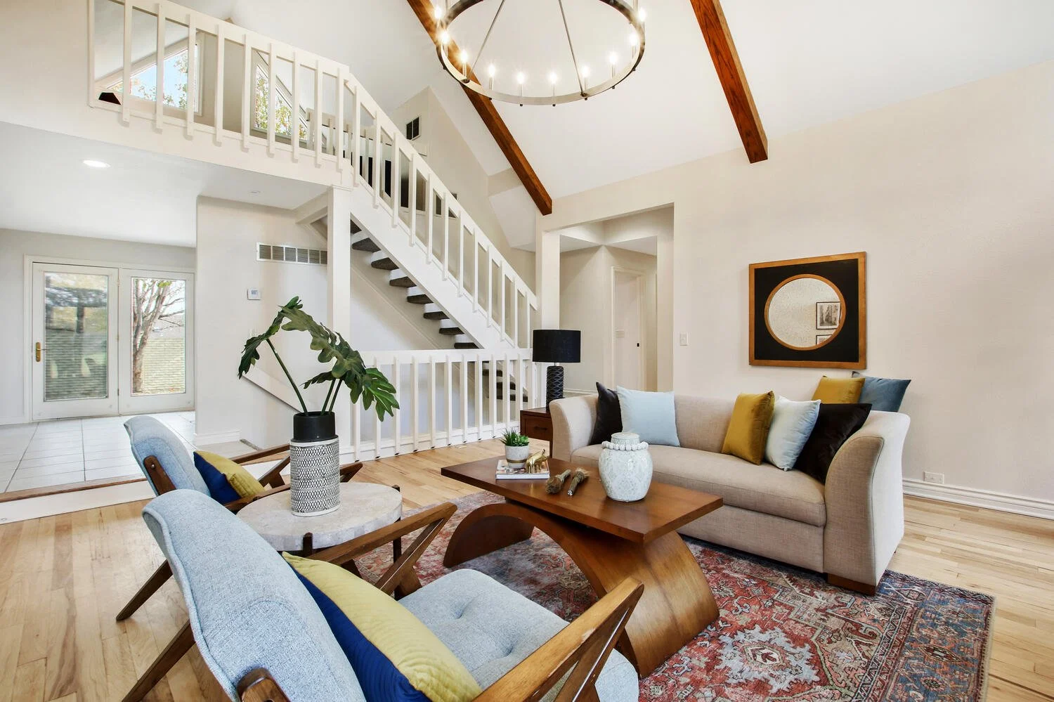 Living room with a cream sofa, colorful pillows, a wooden coffee table, a patterned rug, and a large plant in a vase. There are two light blue chairs with yellow pillows, a black table lamp, and a decorative mirror on the wall. A staircase with white railings and a chandelier are visible.