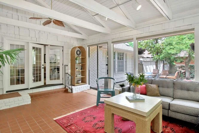 Sunroom with large windows, white brick walls, tiled floor, wooden ceiling, gray couch with pillows, red rug, coffee table with a plant and book, green chairs, and a view of an outdoor backyard with trees and patio furniture.