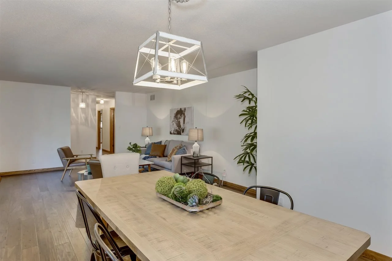 Open-concept living and dining room with wooden dining table, chairs, a sofa, armchair, and decorative plants with a modern chandelier.