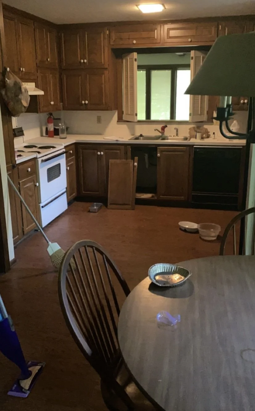 A kitchen with wooden cabinets, a white stove, a window above the sink, and a wooden dining table with chairs. Cleaning supplies and bowls are on the floor.