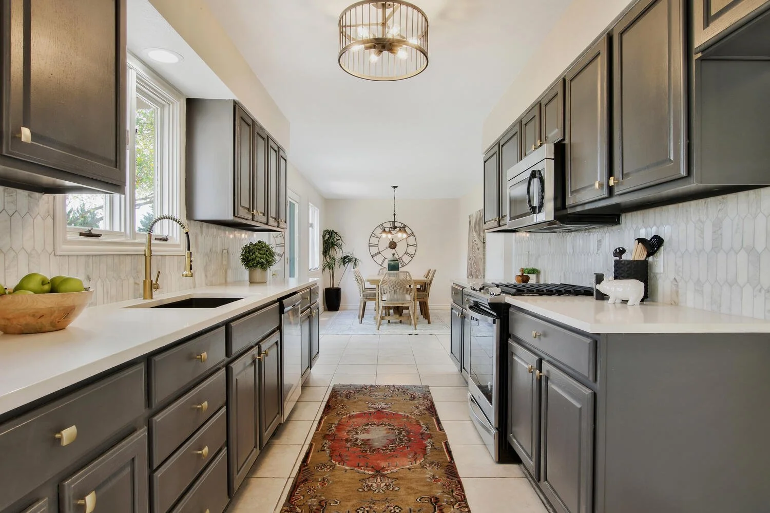 A bright kitchen with gray cabinets, white countertops, a sink, and a window looking outside. There is a decorative rug on the floor, and the kitchen opens into a dining area with a round table, chairs, and a large wall clock.