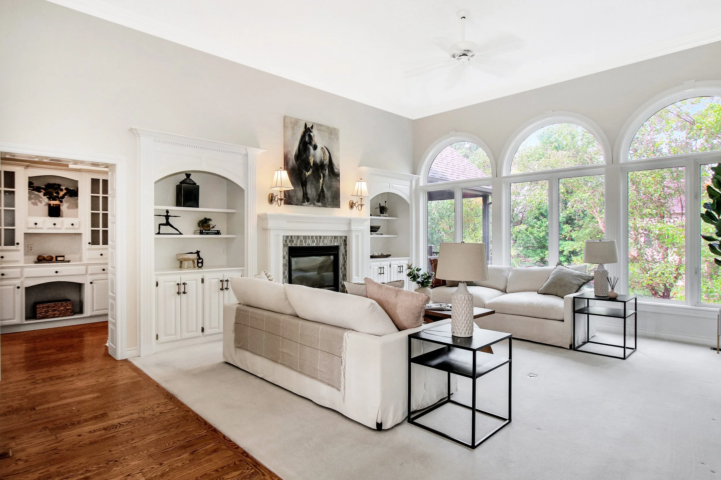 Bright living room with white walls and large arched windows overlooking trees. Contains white sofas, black side tables with lamps, and built-in white shelves.