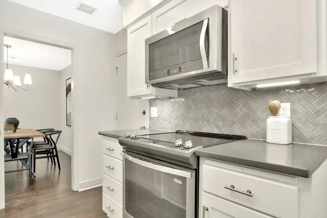 Modern kitchen with white cabinets, gray countertops, stainless steel stove and microwave, decorative tile backsplash, and a dining area with black chairs and a wooden table.