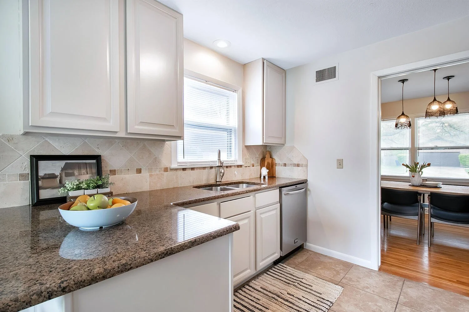 Modern kitchen with granite countertops, white cabinetry, a window above the sink, and a view into a dining room with pendant lights and wooden flooring.