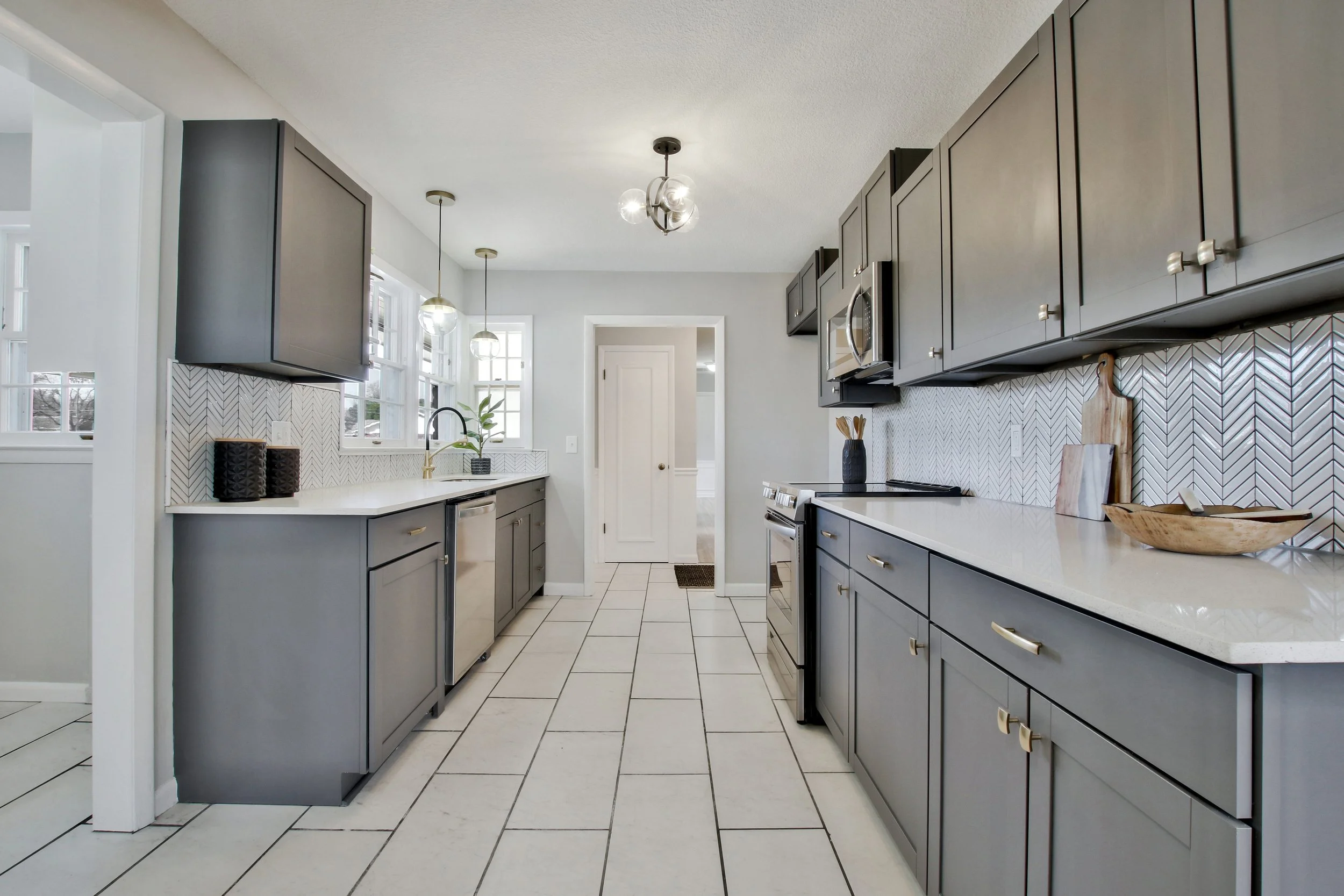 Modern kitchen with gray cabinets, white countertops, and a herringbone-patterned backsplash; includes appliances, pendant lighting, and decorative kitchen items.