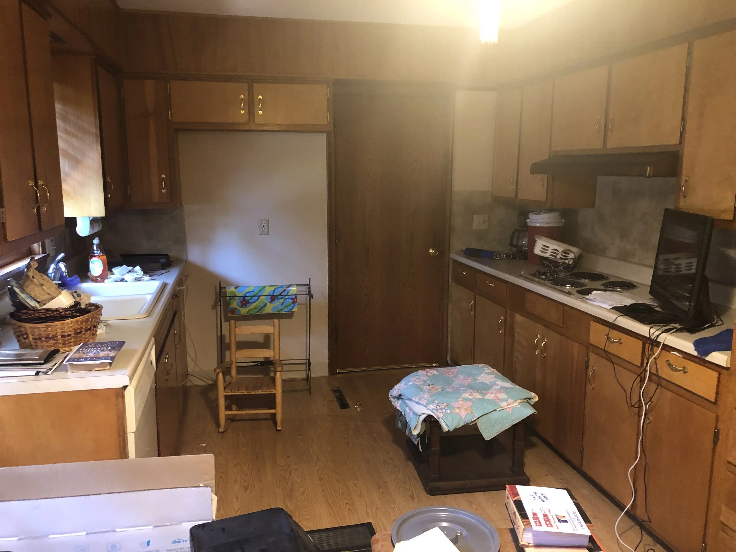 A kitchen with wooden cabinets and a white countertop, a small table with a quilted cloth, a TV on the counter, a laundry drying rack, a bottle of dish soap near the sink, and various kitchen items scattered around.