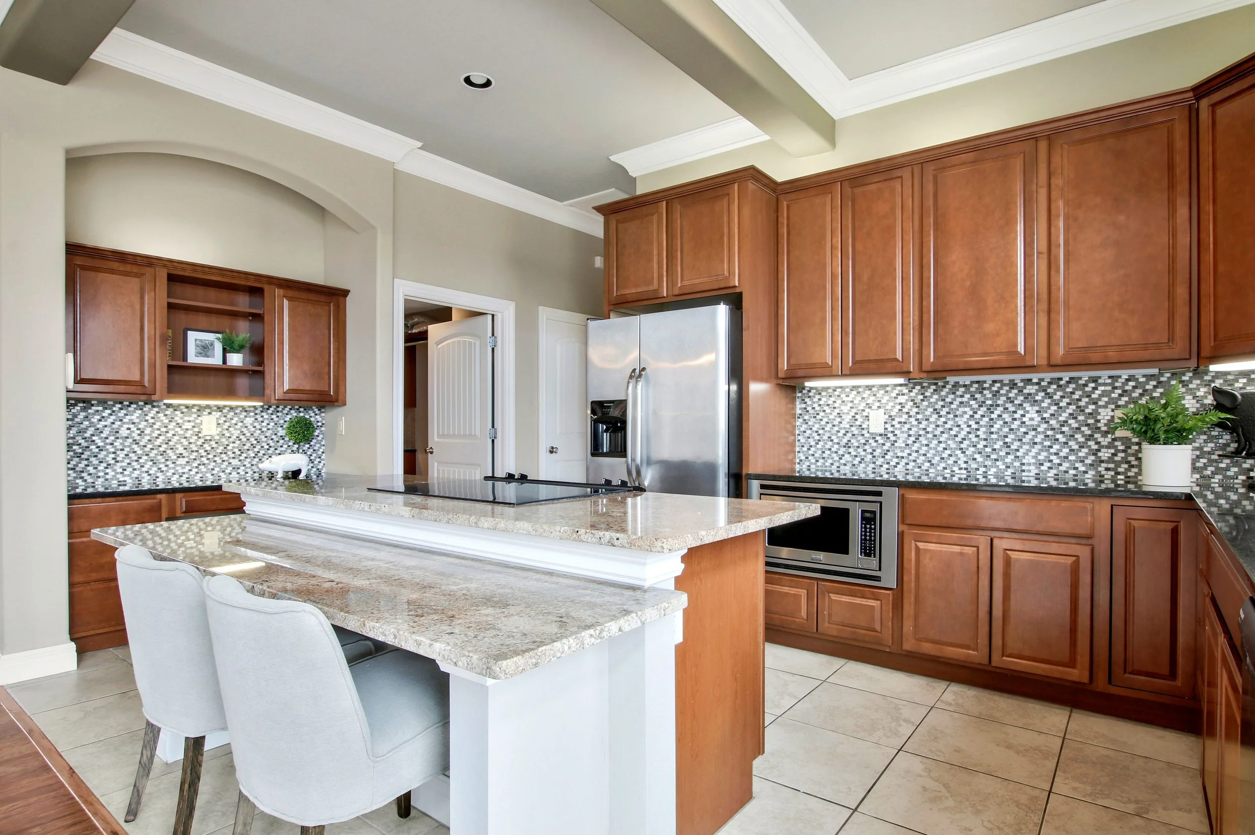 Modern kitchen with wooden cabinets, granite countertops, stainless steel appliances, mosaic tile backsplash, and a small dining table with two chairs.