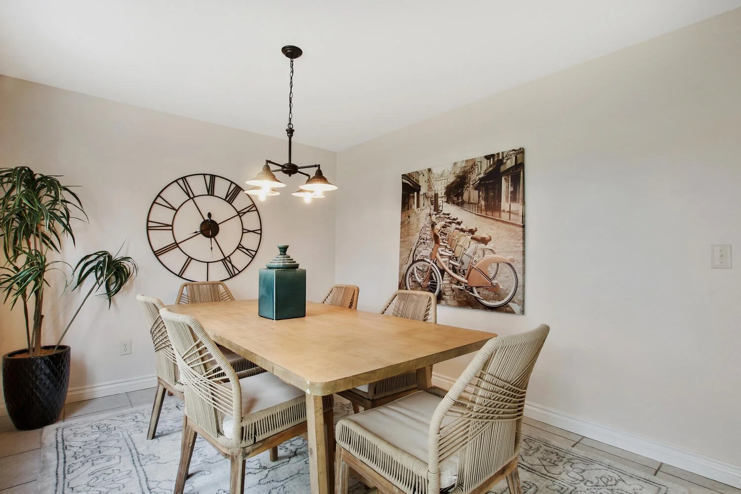 A dining room featuring a wooden table with six rattan chairs, a large wall clock, a potted plant, a framed black and white cityscape photograph, and a three-bulb chandelier
