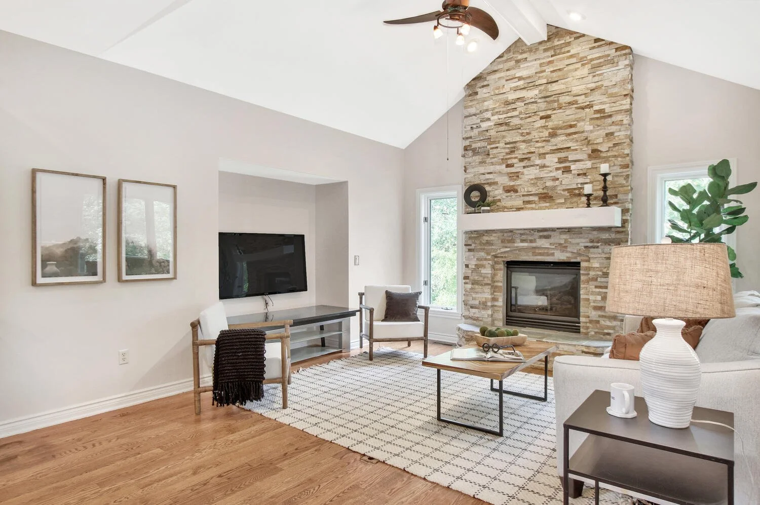 Living room with a stone fireplace, white walls, a ceiling fan, a flat-screen TV, and seating including white armchairs and a sofa, with natural light from windows.