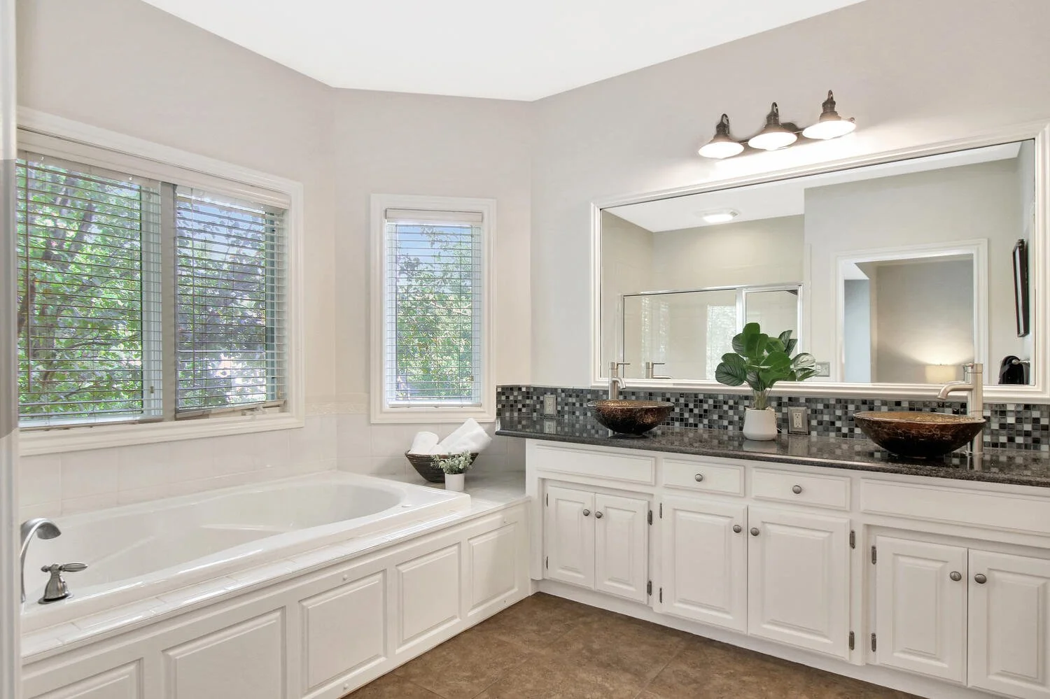 Bright bathroom with a white bathtub, windows with blinds, and a double vanity with vessel sinks and a large mirror.