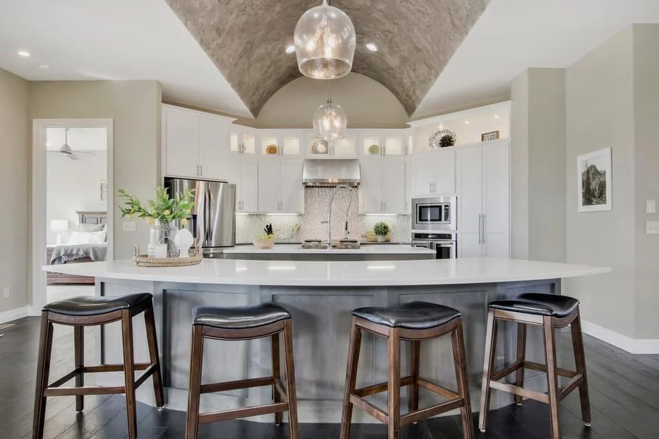 Modern kitchen with white cabinets, stainless steel appliances, a large island with a white countertop, four wooden stools with black cushions, and three pendant lights hanging from a vaulted ceiling.