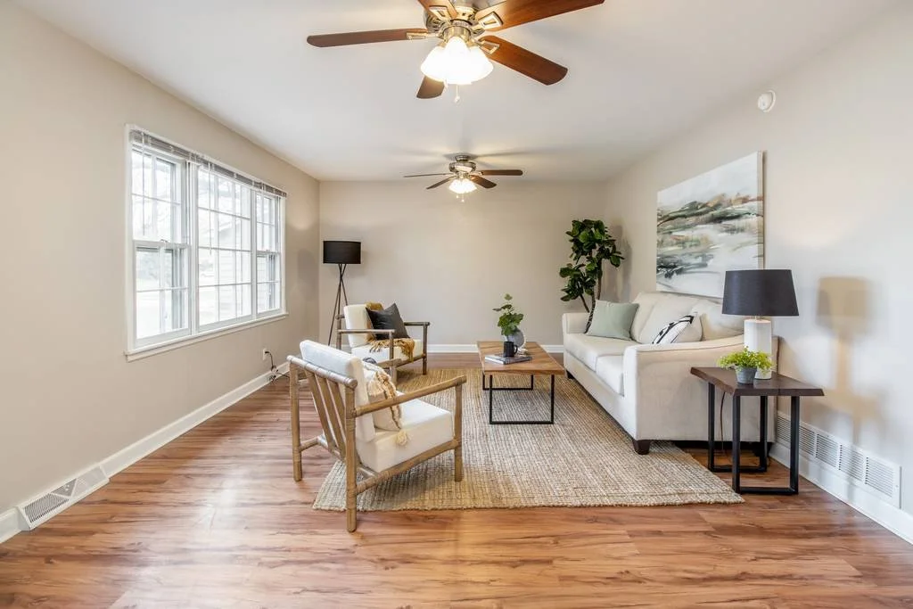 Living room with beige walls, hardwood floors, a white sofa with pillows, two armchairs, a wooden coffee table, a side table with a black lamp, a potted plant, a large landscape painting, a window with blinds, and ceiling fans with lights.