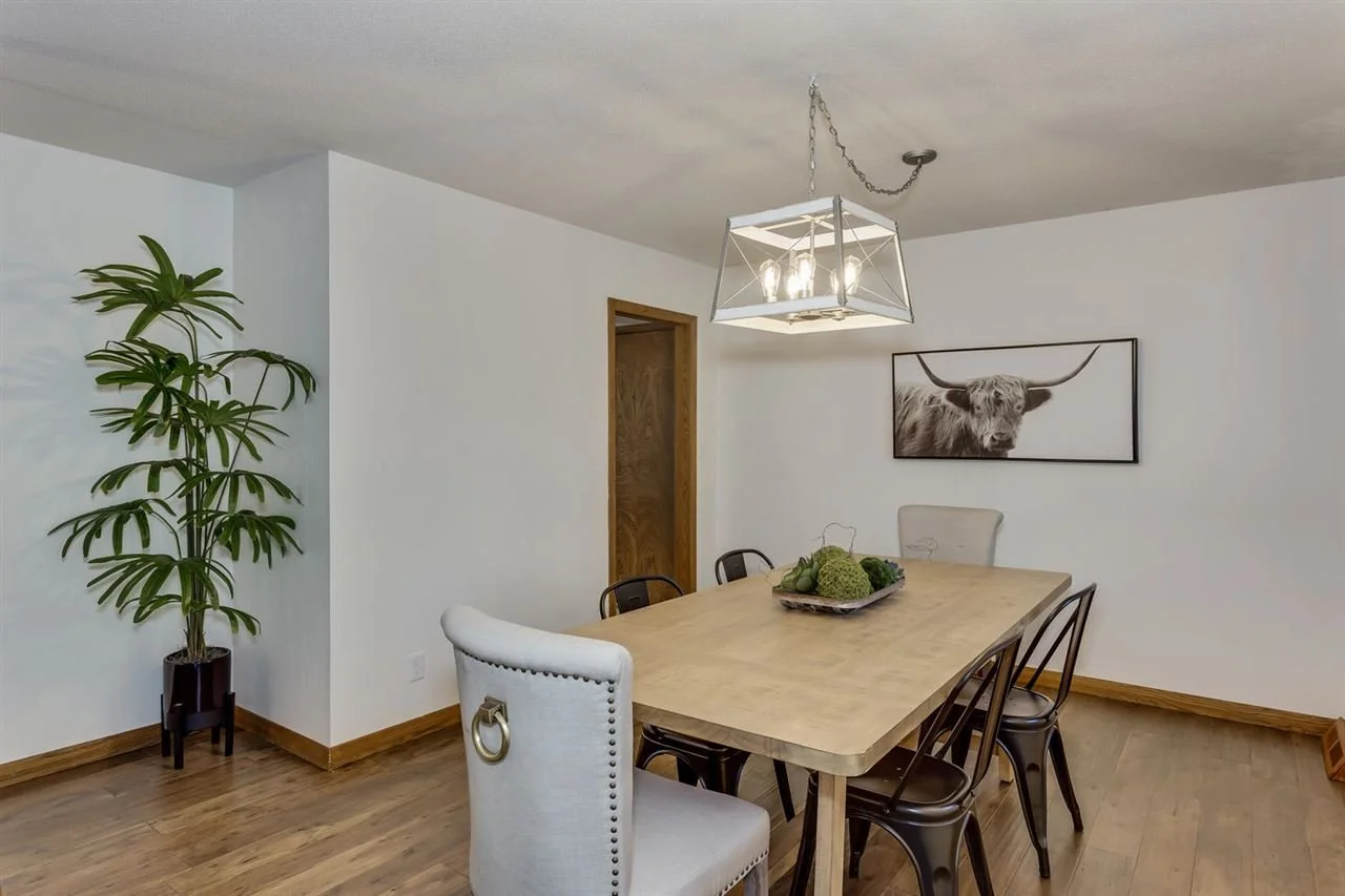 A dining room with a wooden table, six chairs, a large potted plant, artwork of a highland cow on the wall, and a modern chandelier light fixture.