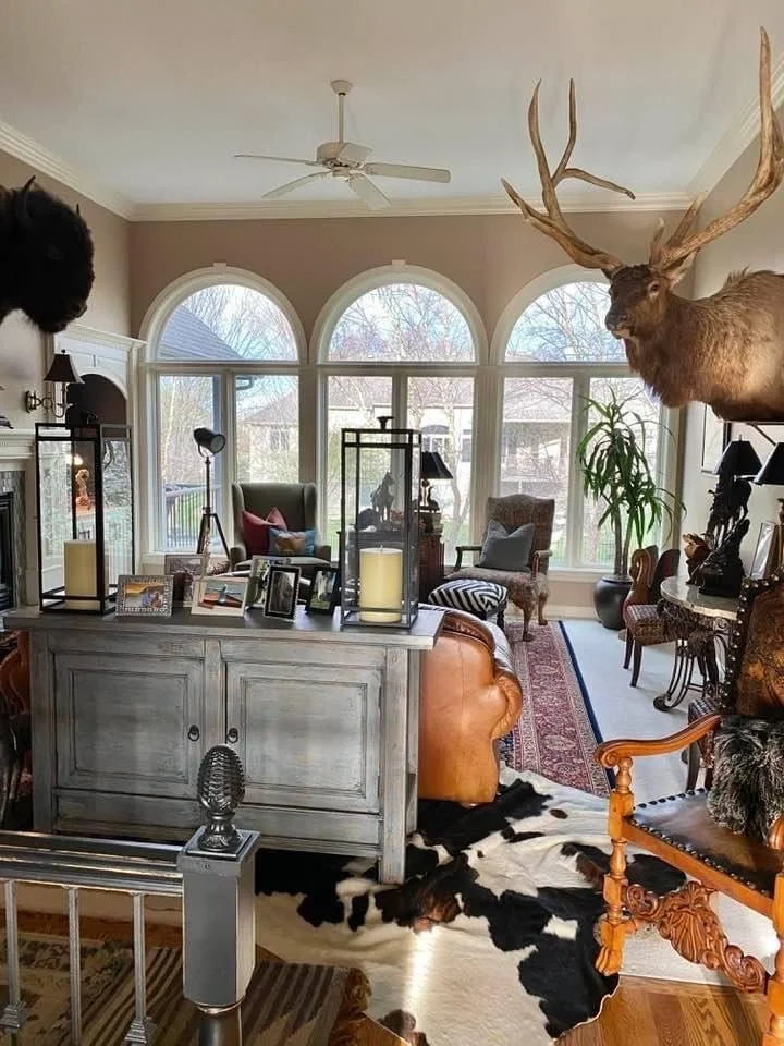 Living room with large arched windows, a mounted deer head with antlers on the wall, a cowhide rug, a white cabinet with framed photos, a wooden armchair, and various decorative items.
