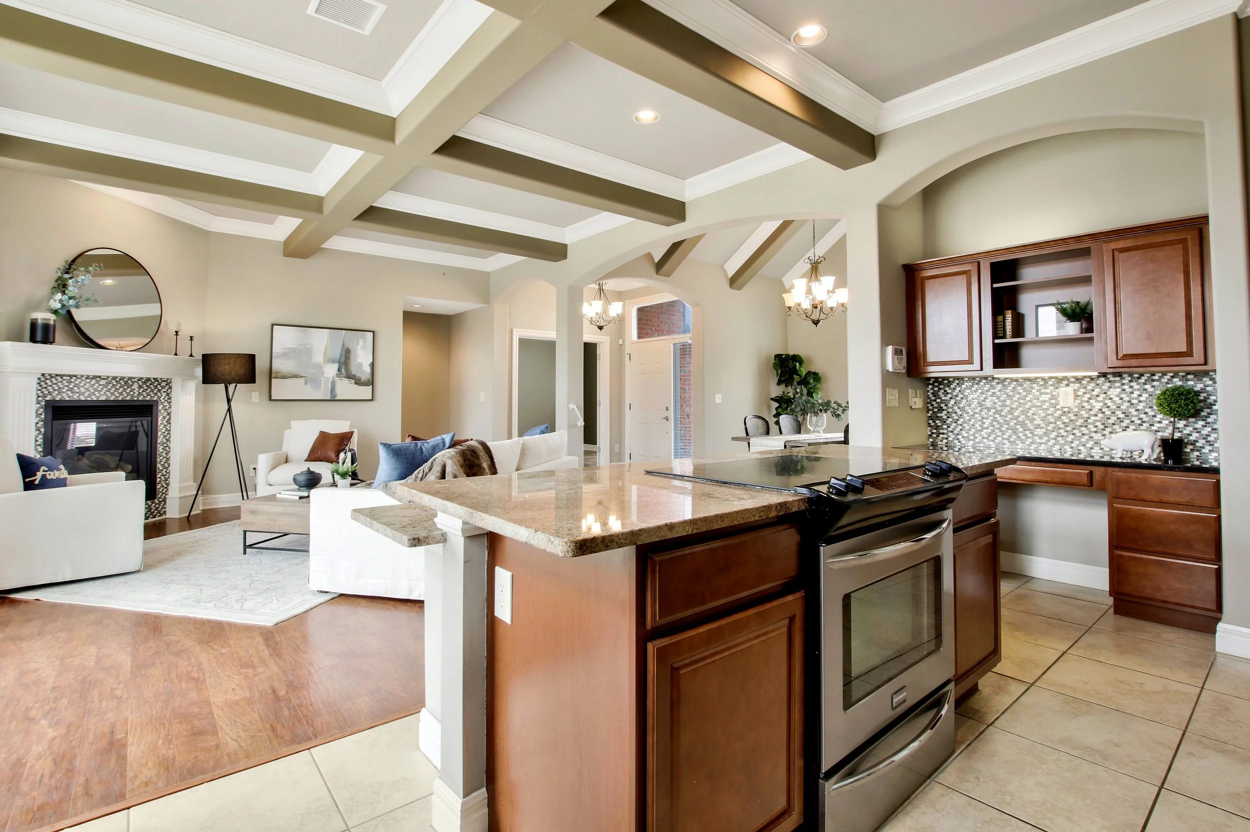 Open-concept living room and kitchen in a modern home with beige walls, wooden cabinets, a granite countertop, stainless steel stove, and decorative ceiling beams.