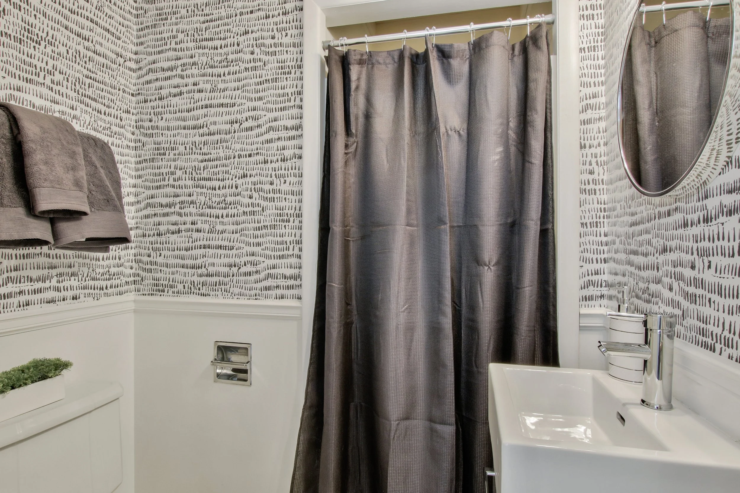A bathroom with patterned wallpaper, a gray shower curtain, a white sink, a round mirror, gray towels on a wall-mounted rack, and a small green plant on a white cabinet.