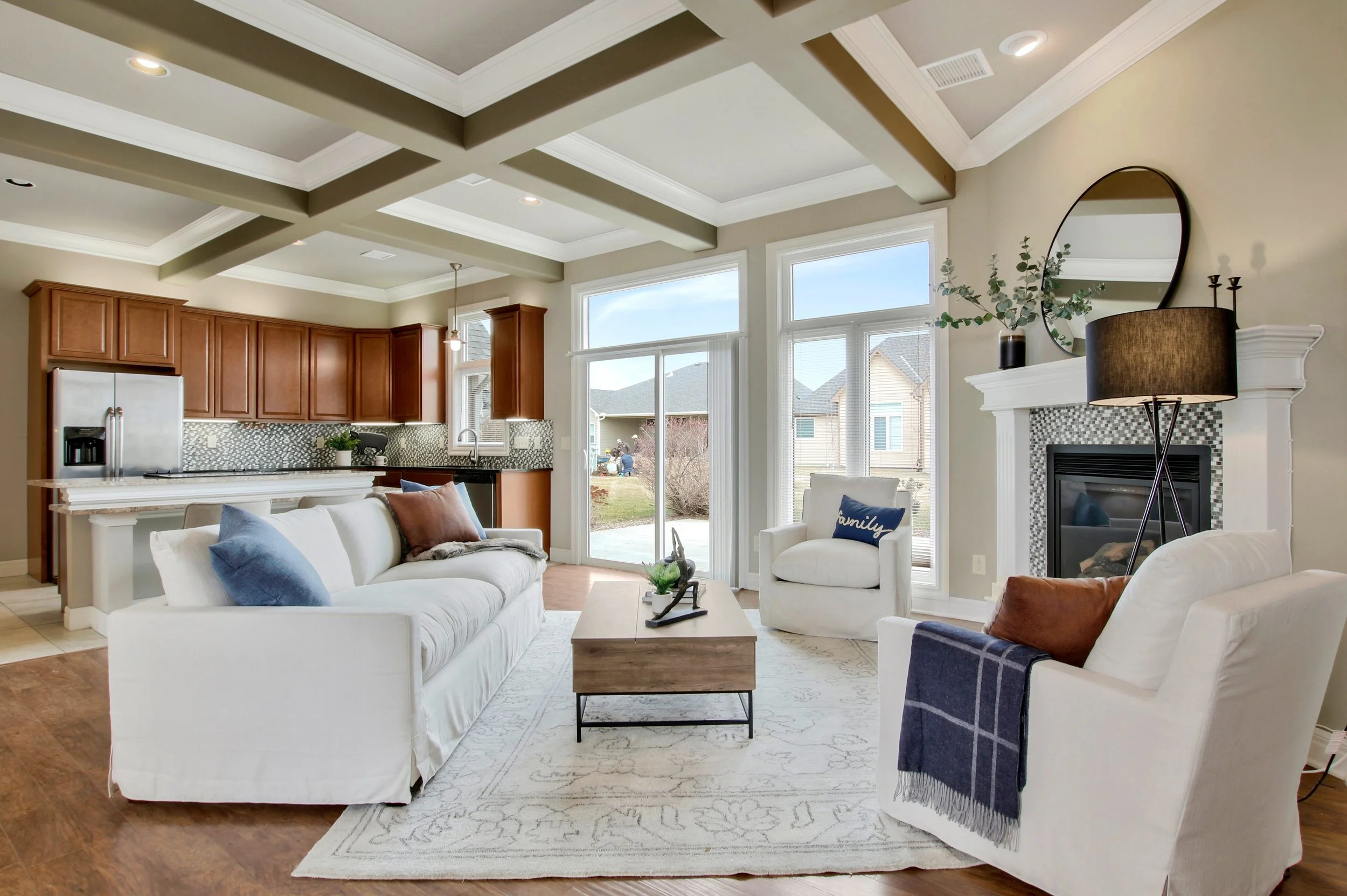 A cozy living room with white sofas and armchairs, a wooden coffee table, a fireplace with a round mirror and decorative items on the mantle, large windows letting in natural light, a ceiling with exposed beams, and an open kitchen in the background with wooden cabinets and a mosaic tile backsplash.