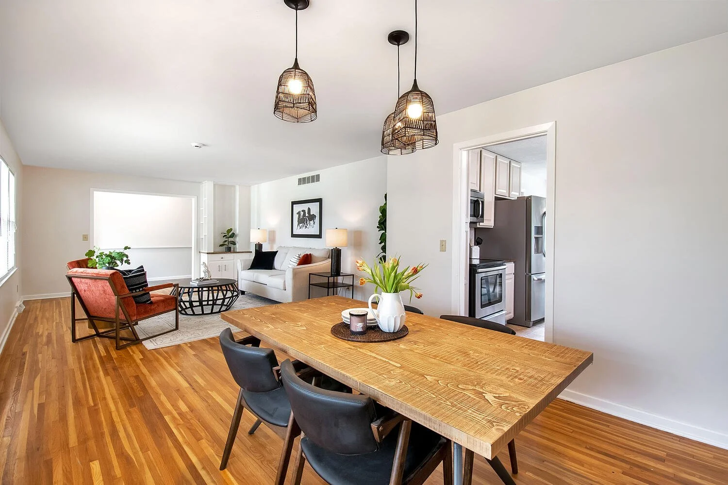 Open-concept living and dining area with hardwood floors, white walls, and modern black light fixtures. The living room has a white sofa, black and orange throw pillows, two side tables with lamps, and a red armchair near a window. The dining area features a wooden table with black chairs, a white vase with flowers, and a small tray with candles. The kitchen with stainless steel appliances is visible through an open doorway.