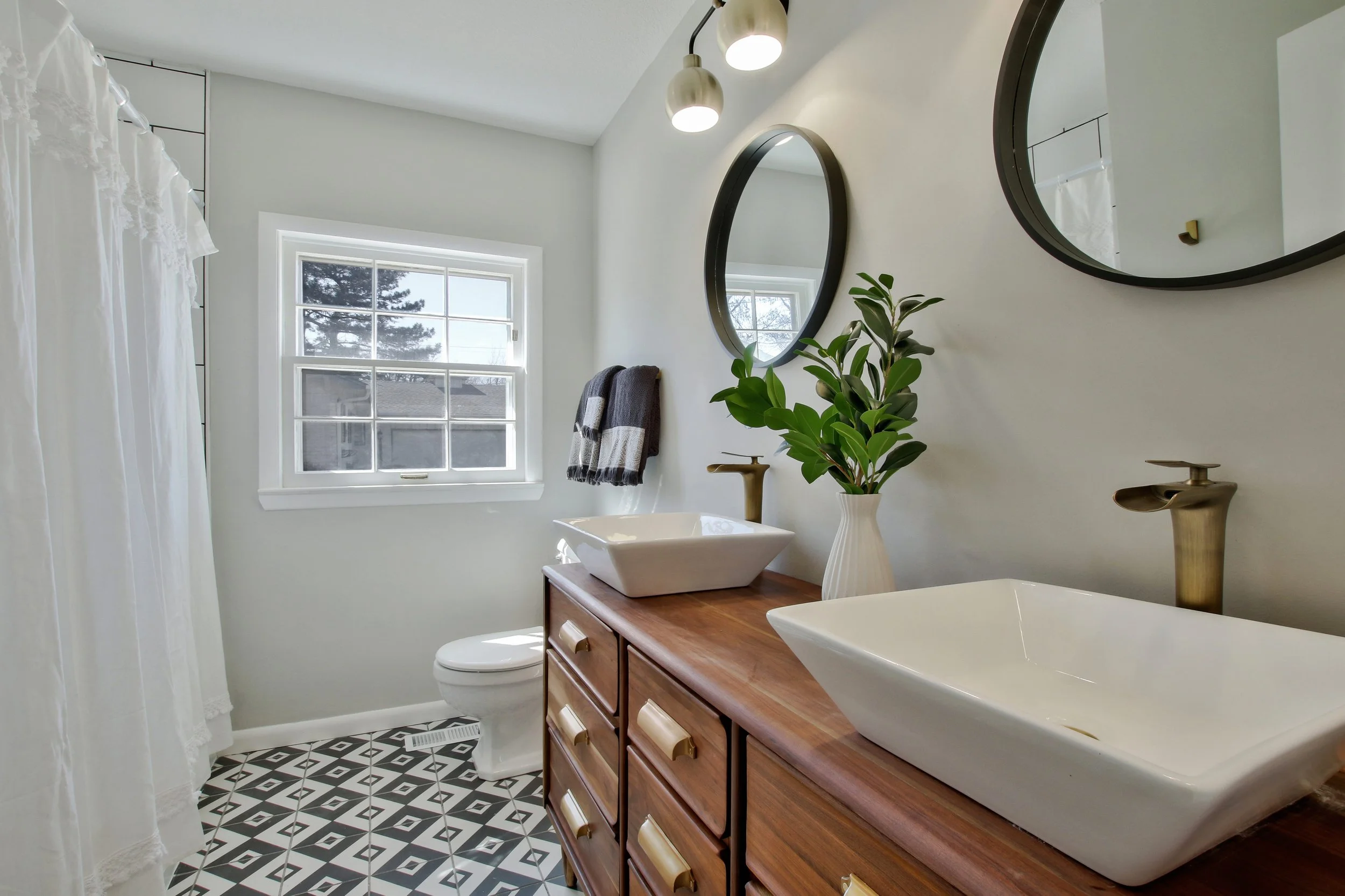 Modern bathroom with double vessel sinks on a wooden vanity, black and white geometric floor tiles, a window, circular mirrors, a brass faucet, a plant in a white vase, and a white shower curtain.