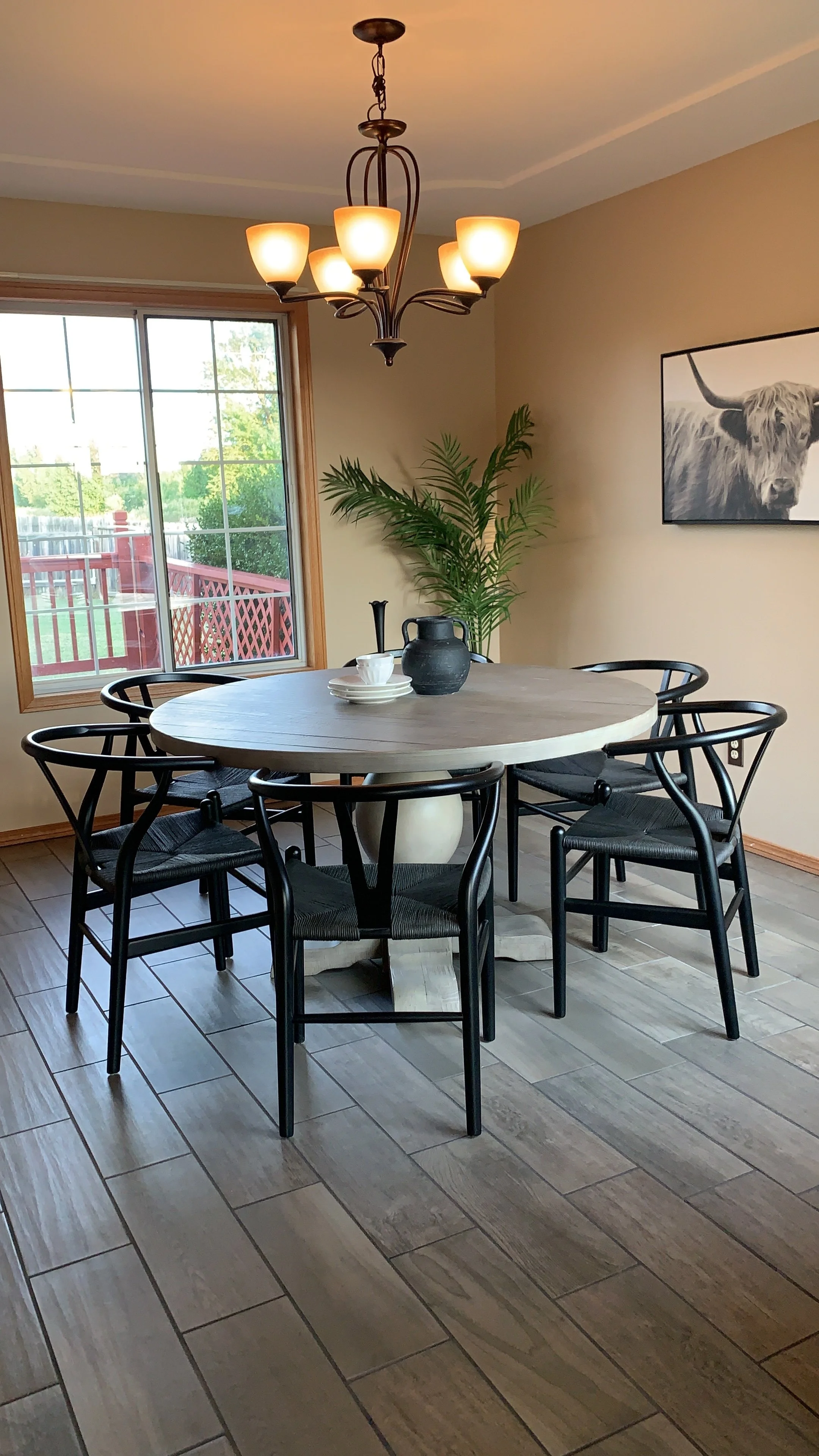 A dining room with a round wooden table, six black chairs, a chandelier light fixture, a large window with a view of a red deck and green trees, a potted plant, a black vase, and a picture of a highland cow on the wall.