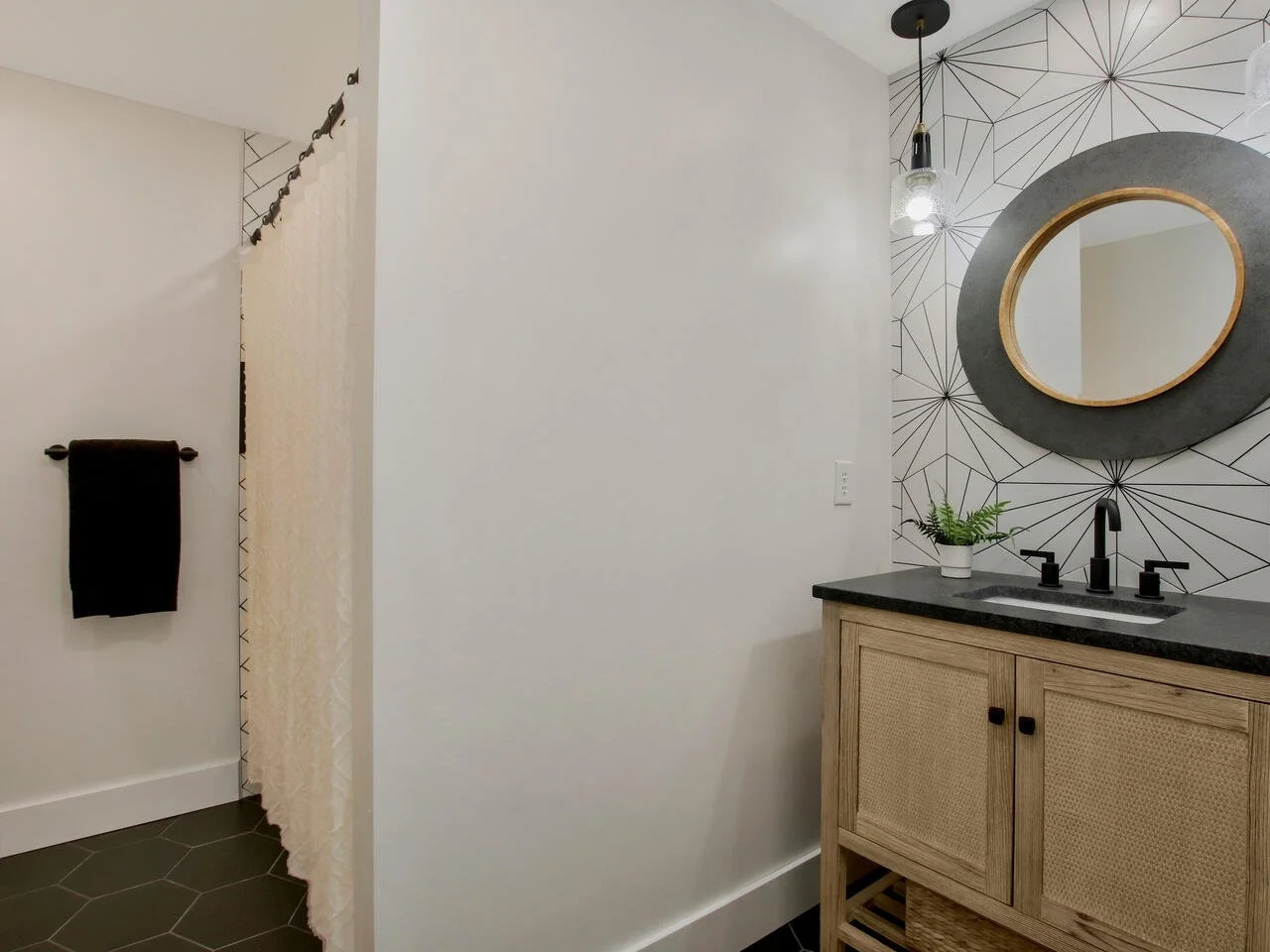 Bathroom with geometric patterned wallpaper, a round mirror with a black frame, a wooden vanity with black hardware, a black faucet, a potted plant, and a light hanging from the ceiling. The room has dark hexagonal floor tiles and a towel on a black towel bar.