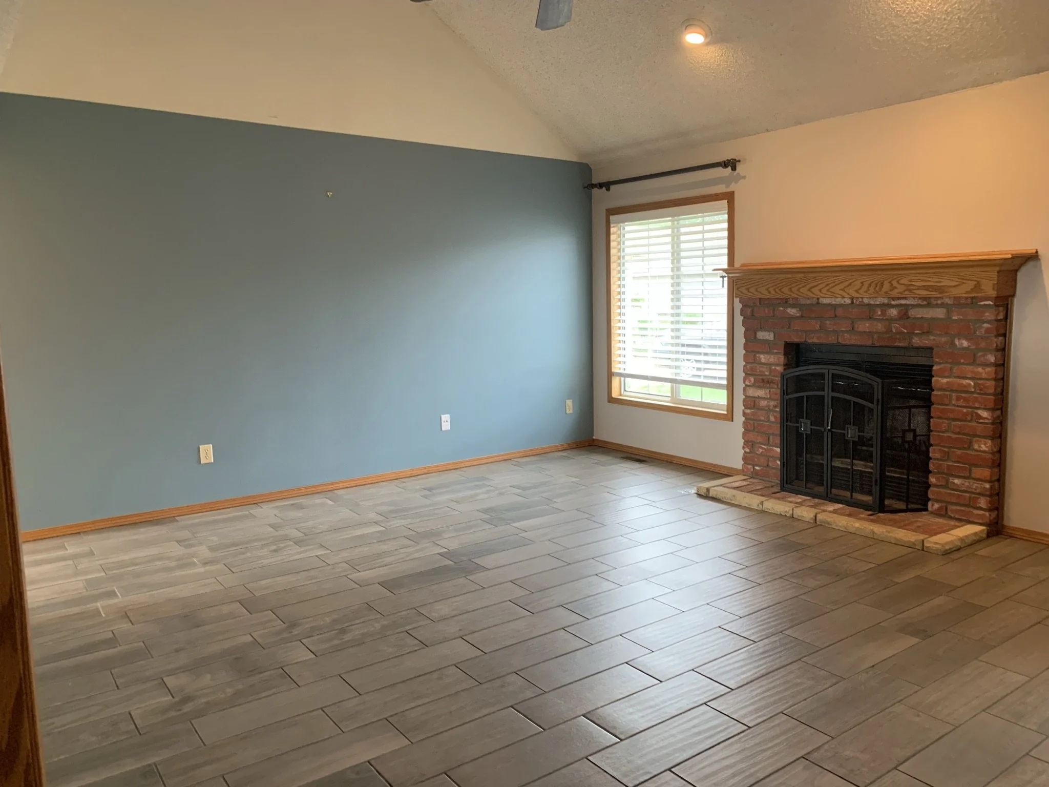 Empty living room with blue wall, brick fireplace, window with blinds, wood trim, and tile floor.