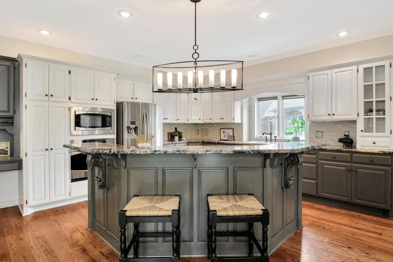 Modern kitchen with white upper cabinets, gray lower cabinets, a large central island with a granite countertop, two wooden stools, a black chandelier with candles, stainless steel appliances, and a window above the sink.