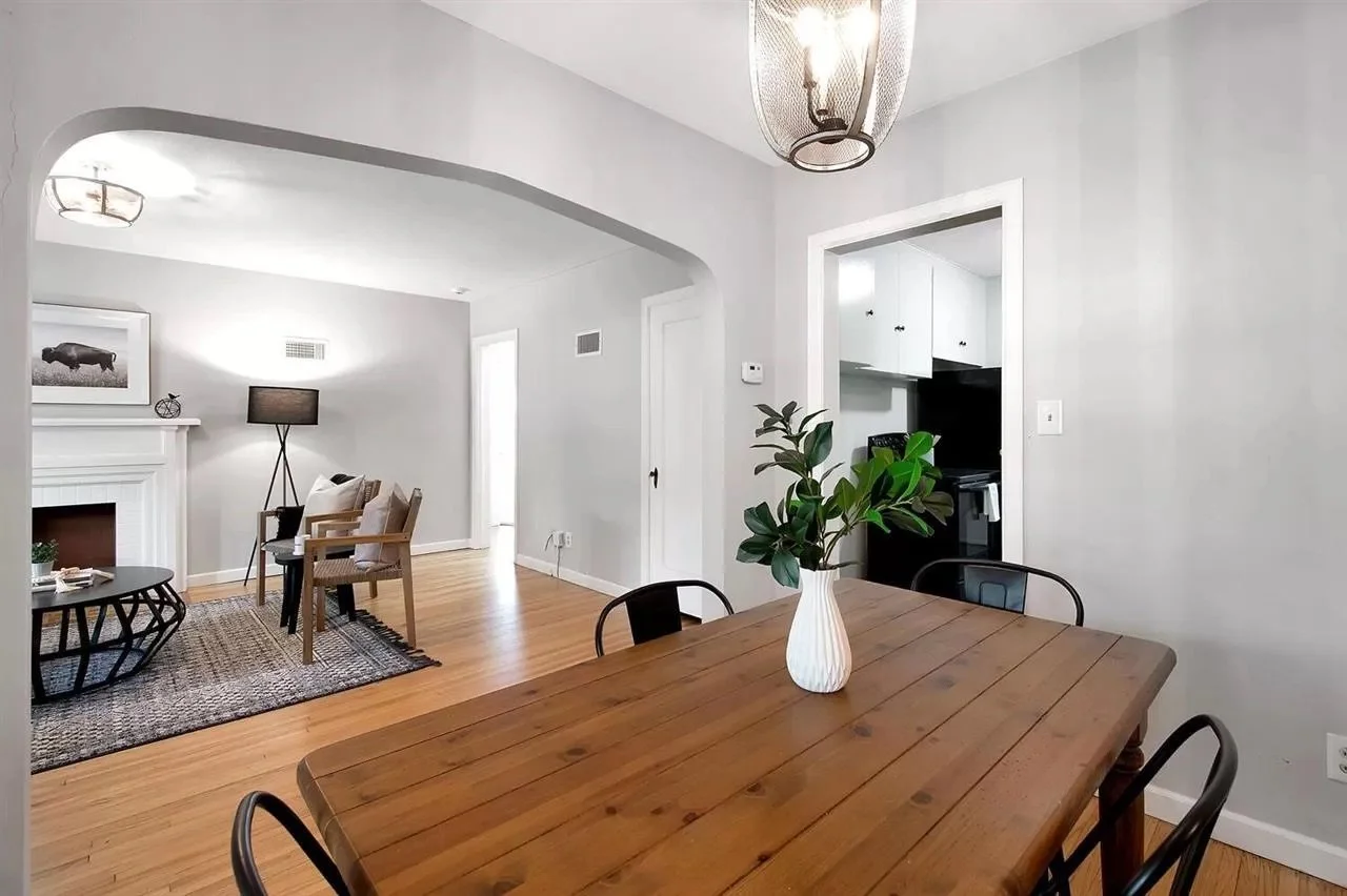 Living and dining room with light gray walls, wooden floors, a wooden dining table with a white vase and green leaves, and a living area with chairs, a black lamp, and a fireplace.