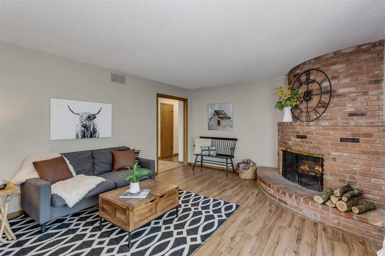 Living room with gray sofa, brown pillows, black and white patterned rug, wooden coffee table, fireplace with logs, wall clock, and wall art, hardwood floors, beige walls.