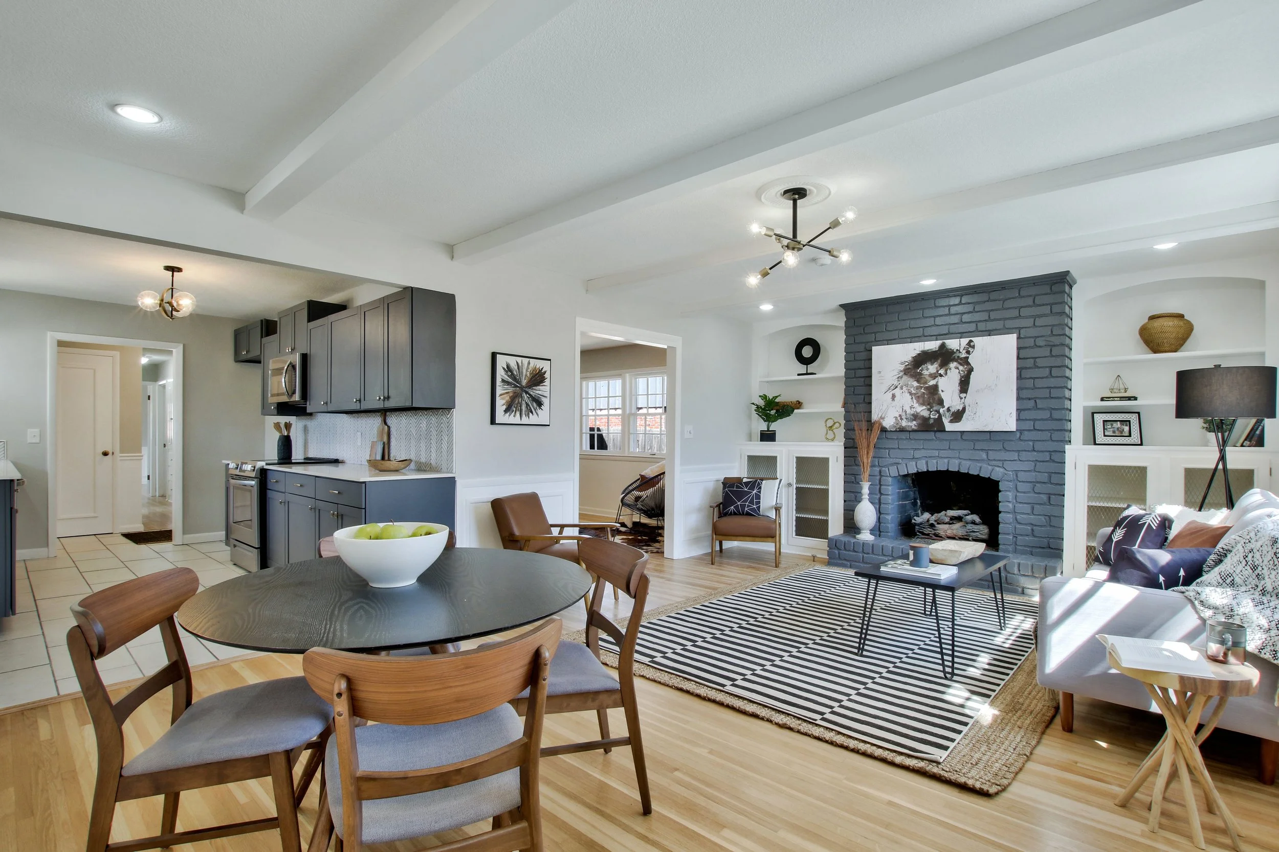 Living room with black brick fireplace, white built-in shelves, striped rug, white sofa with pillows, wooden coffee table, and a dining area with a round wooden table and four chairs. Kitchen in background with dark cabinets and stainless steel appliances.
