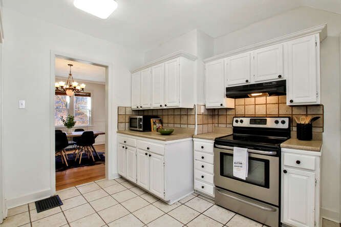 Kitchen with white cabinets, beige tiled backsplash, and a stainless steel stove. There is a microwave on the counter and a black utensil holder. Through the doorway, there is a dining room with a chandelier and wooden table with chairs.
