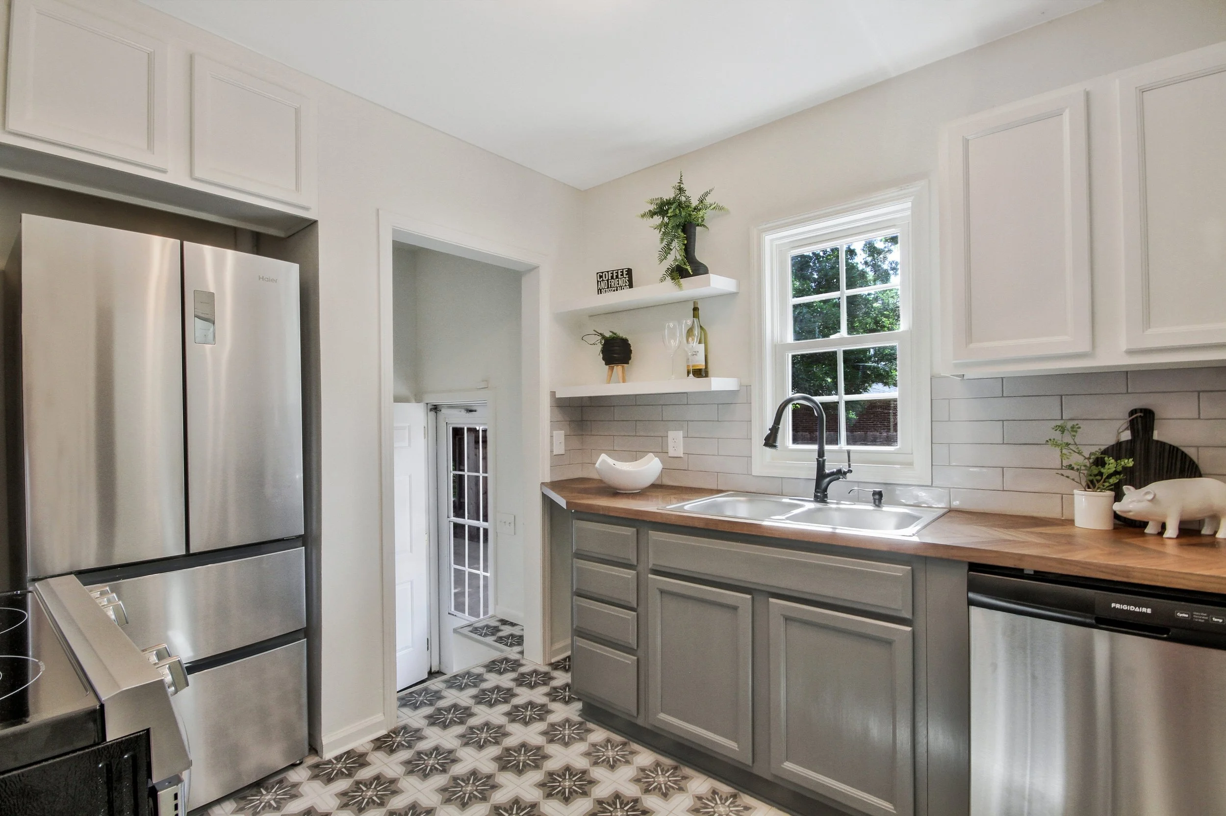 Bright kitchen with gray cabinets, wooden countertop, stainless steel refrigerator, and patterned tile floor, decorated with small plants and kitchenware.