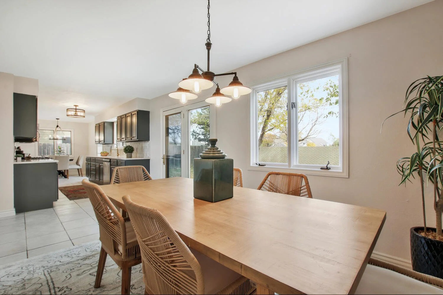 Modern dining room with a wooden table, wicker chairs, and a hanging light fixture, adjacent to a kitchen with dark cabinets and large windows showing outdoor trees.