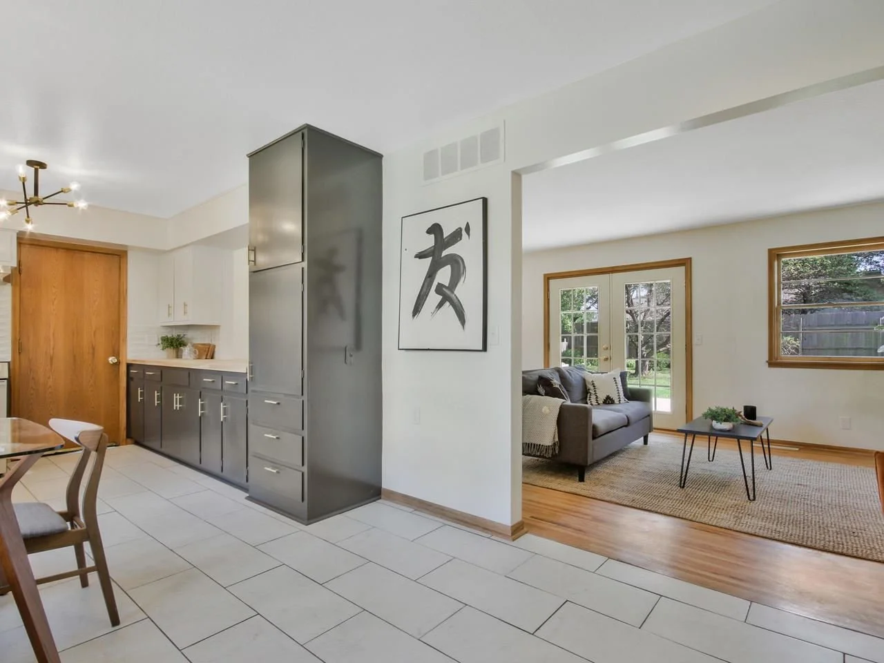 Open-concept living space with a kitchen and sitting area, featuring light-colored tiled floors, wooden cabinets, and a modern chandelier in the kitchen; the sitting area has a gray sofa, large windows, and glass doors leading outside, with a rug and small coffee table.