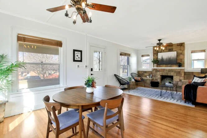 Living room with a dining area, hardwood floors, large windows, a ceiling fan, a stone fireplace with a mounted TV, and modern furniture.