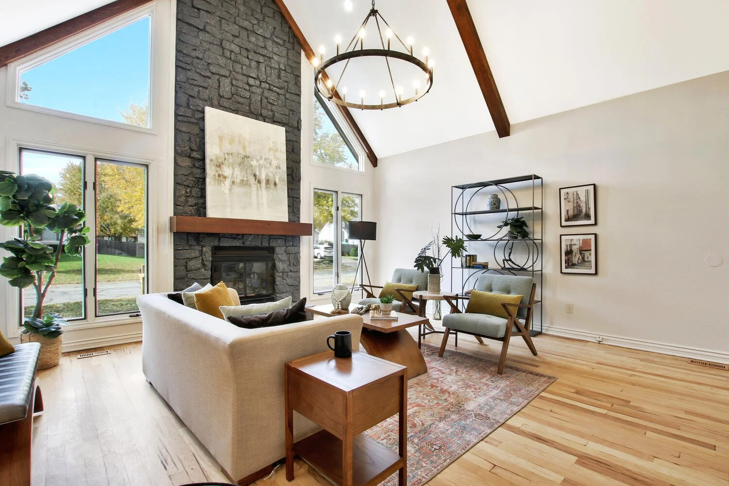 Living room with large windows, a stone fireplace with a wooden mantel, and modern furniture including a white couch, armchairs, and a coffee table. Decor includes a tall bookshelf, artwork on the walls, and a black chandelier hanging from a vaulted ceiling with wooden beams.