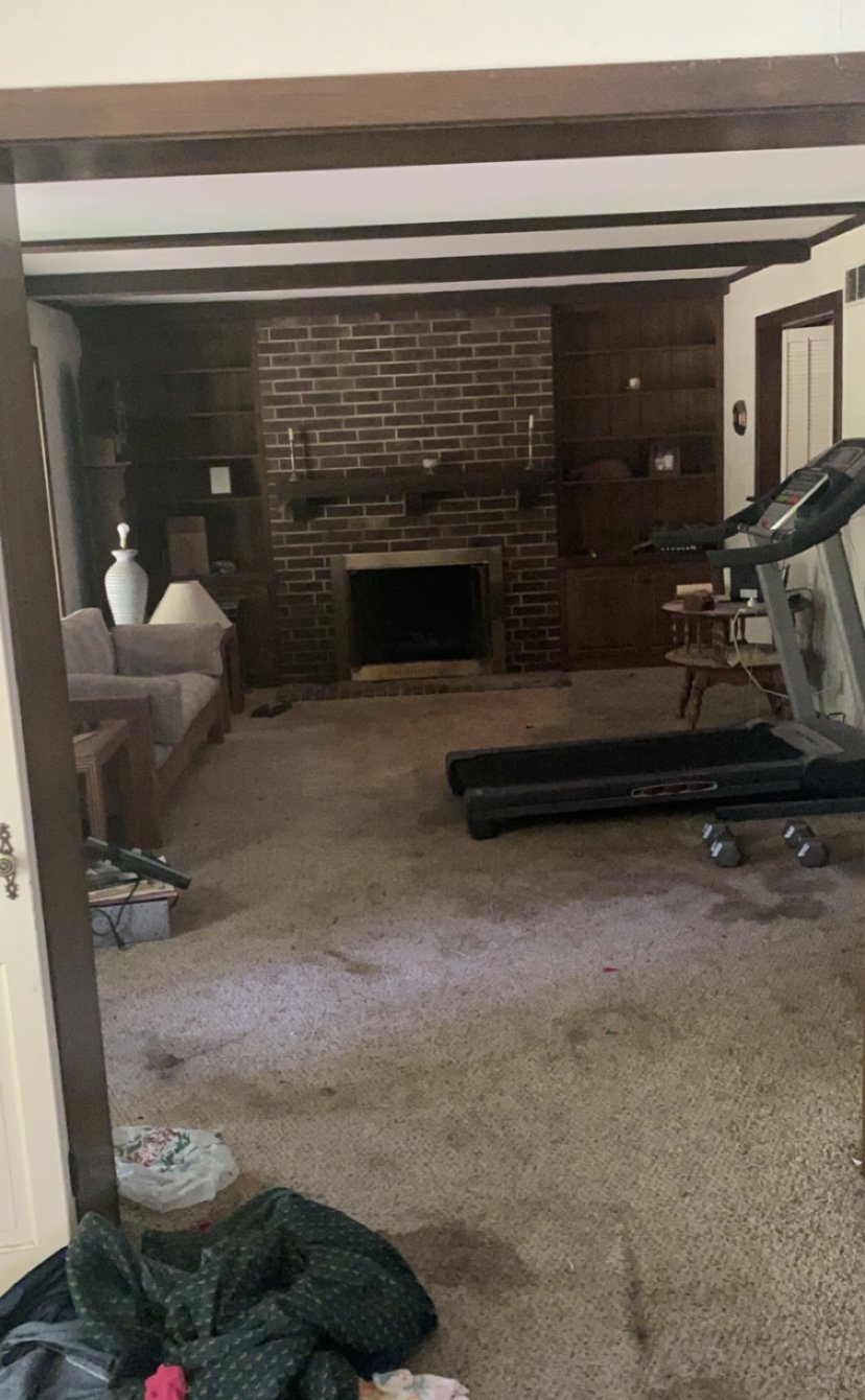 Living room with brick fireplace, treadmill, beige carpet, sofa, table, and wooden built-in shelves.