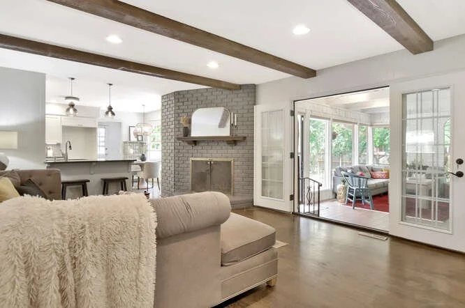 Living room with beige sofa, white walls, exposed ceiling beams, brick fireplace, and large glass doors leading to a sunroom with outdoor seating.