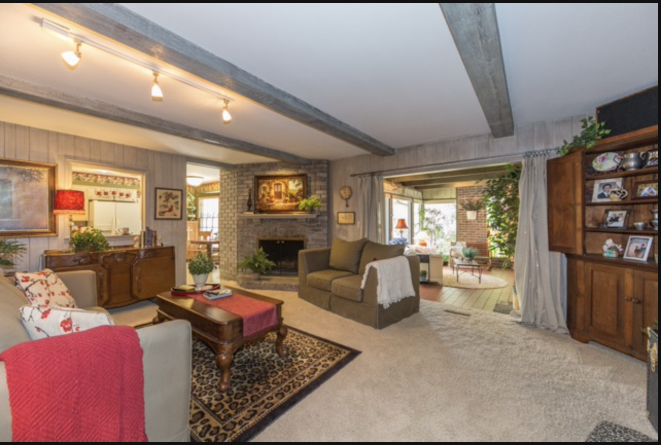 Cozy living room with beige carpet, gray sofa, and brick fireplace, decorated with plants, framed artwork, and wooden furniture, opening into a sunlit sunroom with large windows and greenery.